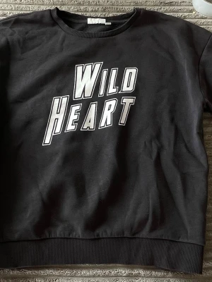 Svart sweatshirt Wild Heart print - Cool svart sweatshirt med stort vitt tryck 'Wild Heart' på bröstet. Tröjan har rund halsringning och ribbade muddar vid ärmslut och nederkant. Perfekt för dig som gillar statement-plagg och streetstyle. Materialet känns mjukt och bekvämt, troligen bomull eller bomullsblandning.