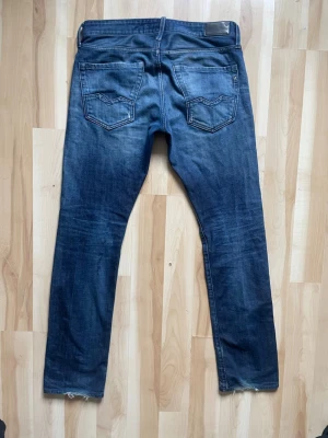 Blå slim fit jeans från Replay - Säljer ett par blå slim fit jeans från Replay med klassisk femficksdesign och snygga slitningar. Jeansen har normal midja, raka ben och detaljerade bakfickor. Tillverkade i mjuk denim med stretch för extra komfort.