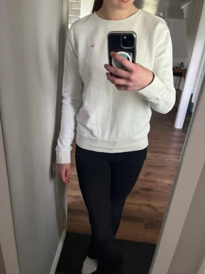Vit sweatshirt från Gant - Vit sweatshirt från Lee med rund halsringning och diskret broderad logga i rosa på bröstet. Tröjan har långa ärmar och muddar vid ärmslut och nederkant. Perfekt för en clean och enkel stil. Tillexempel till ett par jeans eller över en klänning.