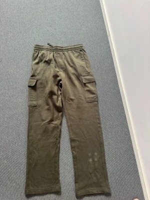 Olivgröna cargobyxor från SOC - Olivgröna cargobyxor från SOC med raka ben och elastisk midja med dragsko. Byxorna har två stora fickor med lock på sidorna och är gjorda i mjukt, sweatshirtliknande material. Perfekta för en avslappnad och streetig stil.