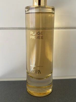 Plage Privée Eau de Parfum Zara - Plage Privée Eau de Parfum från Zara i en elegant, cylindrisk glasflaska med guldiga detaljer och transparent lock. Doften har en ljusgul vätska och flaskan har stilren text i guld. Perfekt för dig som gillar minimalistisk design.