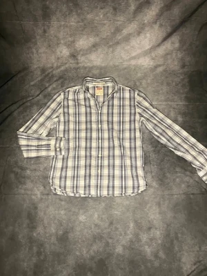 Rutig skjorta från Levi's i blått och vitt - Klassisk rutig skjorta från Levi's i blått, vitt och grått. Skjortan har lång ärm, knappar framtill och en avslappnad passform. Perfekt för dig som gillar tidlös stil och vill ha något som funkar till många olika looks.