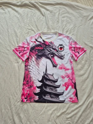 Japan t-shirt med drake och körsbärsblom - Unik t-shirt med korta ärmar och ett stort tryck av en japansk drake, pagod och rosa körsbärsblommor. Färgerna går i vitt, svart och rosa med detaljer i rött. Perfekt för dig som gillar streetwear och asiatiska influenser. Materialet känns som polyester.