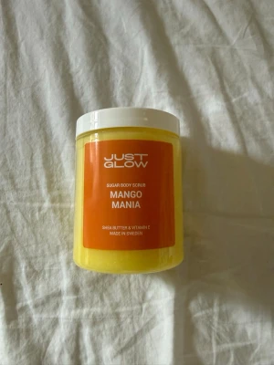 Just Glow Mango Mania Body Scrub - Oöppnad och plomberad. Sockerbaserad body scrub från Just Glow i doften Mango Mania. Kommer i en gul plastburk med vitt lock och orange etikett. Innehåller sheasmör och vitamin E för extra återfuktning. Perfekt för att få len och fräsch hud. Nypris 399kr