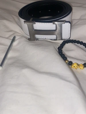 H3rmes bälte - Snyggt vitt skärp med ett stort, silverfärgat H-spänne. Bältet är gjort i skinn och har stilrena svarta sömmar längs kanterna. Perfekt accessoar för att lyfta din outfit med en exklusiv touch. (Bältet är reversibelt som innebär att ens sidan är svart och andra är vit)