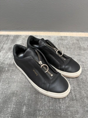 Svarta sneakers från Axel Arigato - Stilrena svarta sneakers från Axel Arigato i mjukt läder med chunky vit sula. Skorna har en unik dragkedja med metallring framtill istället för klassiska snören och diskret guldfärgad logga på sidan. Perfekta för en clean och modern look.