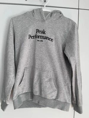 Grå hoodie från Peak Performance - Snygg grå hoodie från Peak Performance med svart broderad logga och text 'Est. 1986' på bröstet. Klassisk känguruficka framtill, ribbade muddar och mjukt material. Perfekt för en avslappnad stil och enkel att matcha med jeans eller joggers.