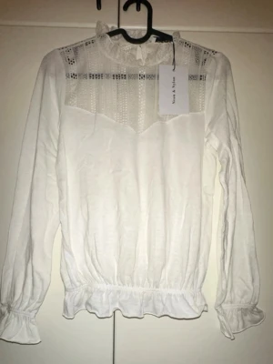 Vit blus med spetsdetaljer och volang - Supersöt vit blus med transparenta spetsdetaljer upptill, hög krage med volang och långa ärmar med resår och volang vid ärmslut. Blusen har resår i midjan för en snygg passform och knäppning bak i nacken. Perfekt för en trendig och romantisk look.