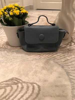 Grå handväska i skinn med handtag - Snygg grå handväska i mjukt skinn med ett stilrent lock och rund knappstängning. Väskan har ett smidigt handtag upptill och detaljerade sömmar. Perfekt storlek för det viktigaste och enkel att matcha till olika outfits. Är öppen med prisförslag😊