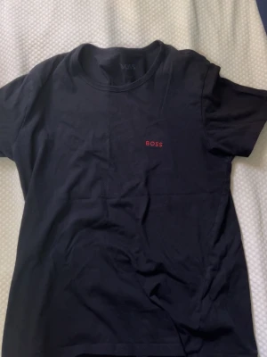 Svart t-shirt från BOSS med röd logga - Snygg svart t-shirt från BOSS med diskret röd logga på bröstet. Klassisk rund hals och korta ärmar. Tillverkad i mjuk bomull som känns skön mot huden. Perfekt för en clean och stilren look.