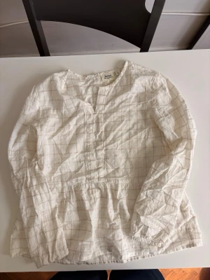 Vit blus med rutmönster från Monki - Säljer en vit blus från Monki med diskret rutmönster i beige. Blusen har långärmad design, rund halsringning med liten slits framtill och är tillverkad i ett lätt och luftigt material. Perfekt för dig som gillar en enkel och stilren look.