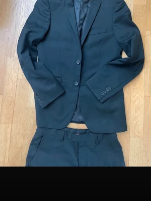 Svart kostym med kavaj och byxor - Säljer en klassisk svart kostym med tvådelad design: kavaj med två knappar och matchande byxor. Kavajen har bröstficka och knappar på ärmsluten. Perfekt för dig som vill ha en stilren och tidlös look.