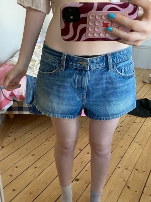 Blå denimshorts med klassisk passform - Säljer ett par blå denimshorts med klassisk femficksdesign och knappstängning. Shortsens passform är normal och de har en midwaist. Perfekta för sommarens alla äventyr, snygga att matcha med t-shirt eller linne.