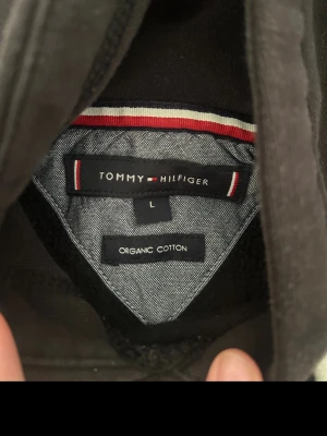 Svart hoodie från Tommy Hilfiger - Svart hoodie från Tommy Hilfiger i ekologisk bomull. Klassisk modell med huva och vita dragsnören, samt broderad logga och text 'EST. 1985' på bröstet. Perfekt för en chill och stilren look.