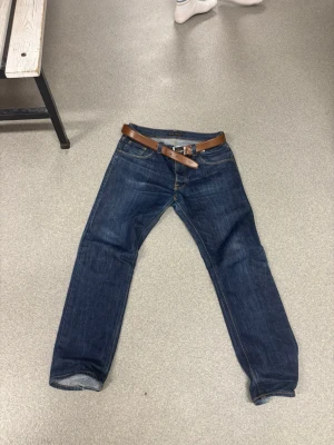 Nudie jeans big Bengt (sällsynt) - Snygga mörkblå jeans i klassisk rak modell med fem fickor och kontrastsömmar. Jeansen har ett brunt läderbälte med silverfärgat spänne. Perfekta för en avslappnad och stilren look. Materialet är denim i bomull.