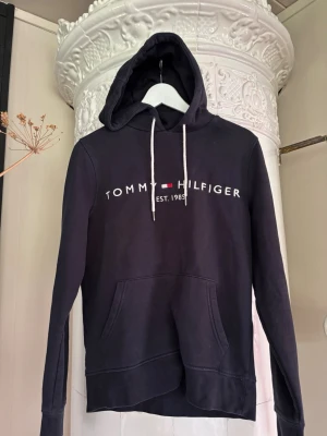 Svart hoodie Tommy Hilfiger XS - Snygg svart hoodie från Tommy Hilfiger i storlek XS. Klassisk design med vit logga och flagga på bröstet, vita dragsnören och stor känguruficka framtill. Tillverkad i mjukt material med borstad insida för extra komfort. Perfekt för en avslappnad och trendig look.