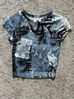 Blå mönstrad croppad topp Bershka - Säljer en blå croppad topp från Bershka med asiatiskt inspirerat mönster. Toppen har korta ärmar och tryck i svart och vitt över hela plagget. Perfekt för en edgy streetstyle-look.