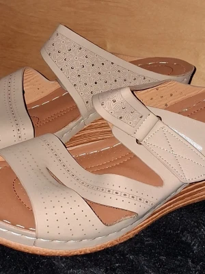 Sandaler med kilklack  - Sandaler med kilklack och dekorativa blommönster på remmarna. Sandalerna har perforerade detaljer och breda remmar i syntetmaterial för extra komfort. Sulan är i en ljusbrun ton med struktur för bra grepp.