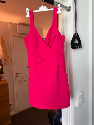 Rosa bandage klänning med breda axelband - Snygg kort rosa klänning med omlottdetalj framtill och breda axelband. Klänningen har en figurnära passform och är perfekt för dig som vill sticka ut. Tillverkad i ett stretchigt material som sitter bekvämt.Helt ny.Storlek märkt som XL men passar storlek L