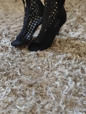 Svarta nätstövlar med klack - Säljer ett par svarta stövlar med öppen tå och klack. Stövlarna har ett coolt nätmönster och snörning baktill som ger en edgy look. Materialet ser ut att vara syntet eller mockaimitation. Perfekta för dig som vill sticka ut.