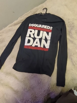 Svart långärmad t-shirt Dsquared2 - ( armarna ser väldigt små ut på bilderna men det är för dom viker sig när man lägger ner tröjan) Cool svart långärmad t-shirt från Dsquared2 med stort vitt och rött tryck på framsidan där det står 'RUN DAN'. Skön passform och tillverkad i mjuk bomull. Perfekt för dig som gillar streetwear och vill sticka ut med ett statement-plagg.