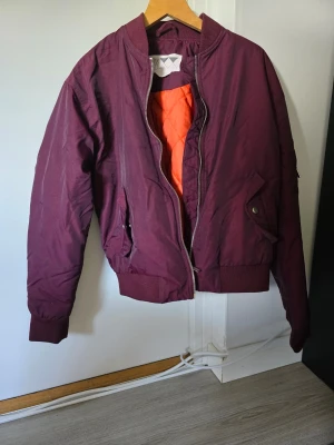 Vinröd bomberjacka med orange foder från junkyard - Snygg vinröd bomberjacka med klassisk passform och orange foder. Jackan har ribbade muddar, dragkedja framtill och två fickor med lock och knapp. Perfekt för dig som vill ha en stilren och trendig look.