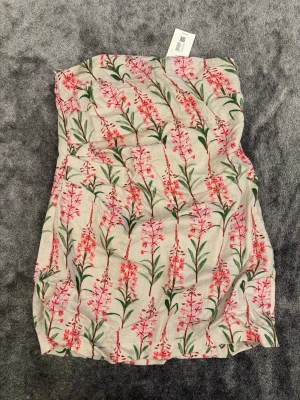 Blommig axelbandslös kortklänning - Söt axelbandslös kortklänning med blommigt mönster i rosa och grönt på beige botten. Perfekt för varma dagar och har en lätt och luftig känsla. Klänningen har en rak passform.