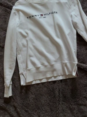 Vit sweatshirt Tommy Hilfiger XS - Vit sweatshirt från Tommy Hilfiger i storlek XS. Tröjan har rund halsringning, långa ärmar och logga broderad framtill med texten 'Tommy Hilfiger EST. 1985'. Tillverkad i mjukt bomullsmaterial och har ribbade muddar vid ärmslut och nederkant. Super snygg o lätt