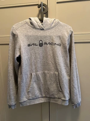 Grå hoodie från Sail Racing - Snygg grå hoodie från Sail Racing med svart logga på bröstet. Klassisk känguruficka framtill och mjuk bomullsblandning. Huva och ribbade muddar vid ärmslut och nederkant gör den extra bekväm. Perfekt för chill dagar eller när du vill ha en clean look.