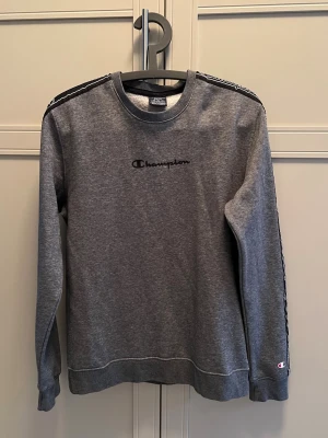 Grå sweatshirt från Champion - Grå sweatshirt från Champion med svart logga broderad på bröstet och svarta band med Champion-tryck längs ärmarna. Klassisk rund halsringning och ribbade muddar vid ärmslut och nederkant. Perfekt för en avslappnad stil.