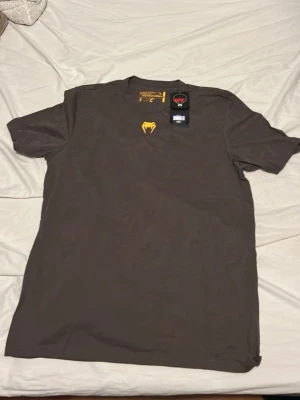 Mörkbrun UFC t-shirt från Venum - Snygg mörkbrun t-shirt från Venum x UFC med gul logga på bröstet. Klassisk passform med korta ärmar och rund halsringning. Perfekt för dig som gillar sportig stil och vill visa ditt intresse för MMA. Tröjan är aldrig använd säljer för att den är för stor
