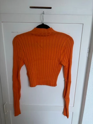Orange ribbad stickad tröja - Trendig orange stickad tröja med ribbad struktur och croppad passform. Tröjan har långa ärmar och en hög hals, perfekt för att addera färg till din outfit. Materialet känns mjukt och stretchigt, vilket gör den bekväm att bära hela dagen.