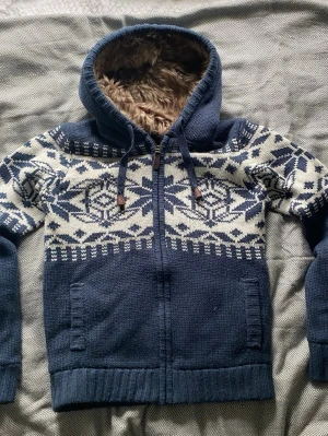 Blå stickad hoodie med mönster - Mysig blå stickad hoodie med vitt nordiskt mönster och dragkedja framtill. Fodrad huva med fuskpäls och dragsko. Två fickor fram och ribbade muddar. Perfekt för kalla dagar och riktigt snygg till jeans.