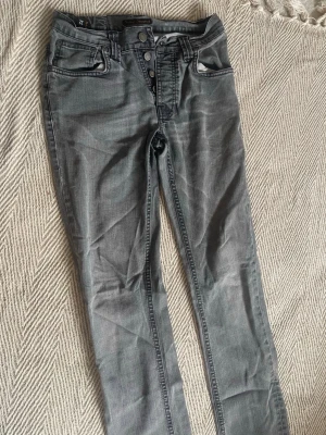 Grå jeans från Nudie Jeans - Snygga grå jeans från Nudie Jeans med klassisk femficksdesign och svarta detaljer på bakfickorna. Jeansen har knappgylf och raka ben. Perfekta för dig som gillar en avslappnad men stilren look.