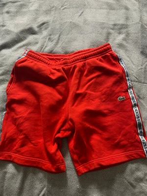 Röda shorts från Lacoste med logoband - Snygga röda shorts från Lacoste med elastisk midja och vita logoband längs sidorna. Klassisk Lacoste-krokodil på vänster ben. Perfekta för sommaren och avslappnade dagar. Mjukt material som känns skönt mot huden.