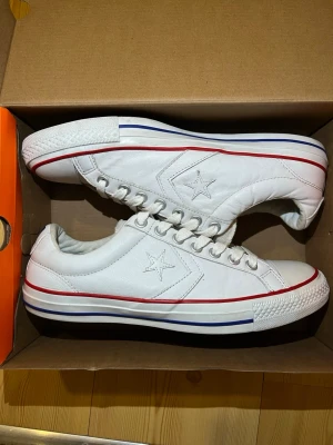Vita Converse One Star sneakers - Säljer ett par klassiska vita Converse One Star sneakers i skinn med snörning. Skorna har röda och blå ränder runt sulan och den ikoniska stjärnan på sidan. Perfekta för dig som vill ha en clean och tidlös look. Kontakta mig för fler bilder.