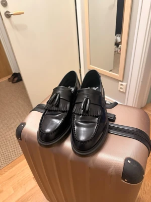 Svarta loafers med tofsar Dr. Martens Adrians Svarta - Snygga svarta loafers från Dr. Martens i blankt läder med klassiska tofsar och fransdetalj framtill. Skorna har en robust sula och stilren design som passar perfekt till både jeans och kostymbyxor. Enbart använd 1 gång.