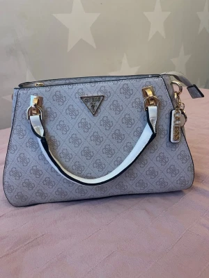 Guess Noelle Luxury Satchel - Snygg beige handväska från Guess med all-over logomönster och guldfärgade detaljer. Väskan har vita handtag med svarta kanter och dekorativa Guess-taggar i metall. Axelrem ingår.