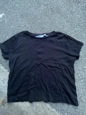 Svart ribbad t-shirt från Fabulous - En enkel svart ribbad t-shirt från Fabulous i storlek S. T-shirten har korta ärmar och rund halsringning, perfekt till jeans eller kjol. Materialet känns mjukt och stretchigt, vilket gör den skön att bära hela dagen.