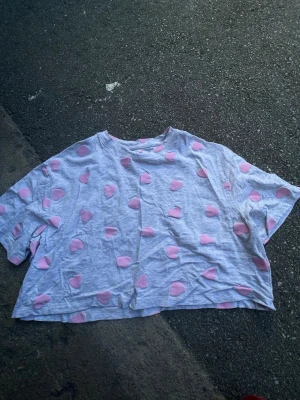 Grå croppad topp med rosa hjärtan - Supersöt croppad topp från Censored i ljusgrått med rosa hjärtmönster över hela. T-shirten har korta ärmar och rund halsringning. Perfekt för dig som gillar gulliga detaljer och en avslappnad stil. 
