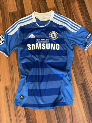 Chelsea Champions League tröja 2012 - Chelsea FC matchtröja från Champions League-finalen 2012 i blått med vita detaljer. Adidas-logga, klubbmärke och Samsung som sponsor på bröstet. Tre vita ränder på axlarna och Champions League-märke på ärmen. 