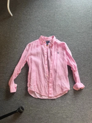 Rosa linneskjorta från Polo Ralph Lauren - Säljer en klassisk rosa skjorta i linne från Polo Ralph Lauren. Skjortan har button-down krage, långa ärmar och den ikoniska blå loggan broderad på bröstet. Perfekt för en avslappnad men ändå stilren look. Den är barn 14-16 år men det blir som en xs.