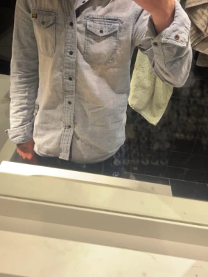 Ljusblå skjorta från Superdry - Säljer en ljusblå skjorta från Superdry med två bröstfickor och knappar framtill. Skjortan har klassisk krage, långa ärmar och en avslappnad passform. Perfekt till jeans eller chinos för en clean look.