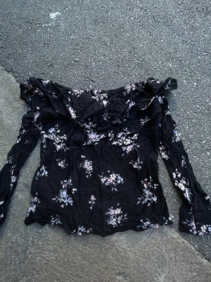Blommig offshoulder blus från H&M Divided - Snygg svart offshoulder blus från H&M Divided med blommigt mönster i vitt och rosa. Blusen har långa ärmar och volangdetalj vid axlarna. Perfekt för dig som gillar romantisk och trendig stil.