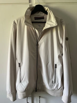 Beige vindjacka från J.Lindeberg - Stilren beige vindjacka från J.Lindeberg med hög krage och hel dragkedja framtill. Två framfickor med dragkedja och diskret logga på ärmen. Jackan har elastiska muddar vid ärmslut och nederkant för en snygg passform. Perfekt för blåsiga dagar.