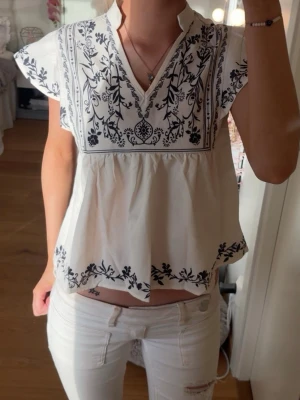Vit blus med svart marinblått broderi - Säljer en luftig vit blus med marinblå broderat blommönster och v-ringning. Blusen har korta ärmar, volangkant nedtill och en avslappnad passform. 