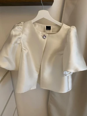 Krämvit puffärm blus Gina Tricot - Stilren krämvit blus från Gina Tricot med puffiga korta ärmar och en dekorativ knapp framtill. Blusen har en lätt glansig satinfinish och rak passform, perfekt för att lyfta vilken outfit som helst. Materialet känns mjukt och exklusivt.