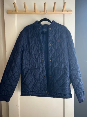 Marinblå quiltad jacka Polo Ralph Lauren - Snygg marinblå quiltad jacka från Polo Ralph Lauren med tryckknappar, ribbad krage och två framfickor. Jackan har det klassiska röda Polo-logot på bröstet och ett rutmönster över hela ytan. Perfekt för kyliga dagar och enkel att matcha.