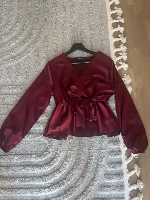 Vinröd omlottblus från Missay - Vinröd omlottblus från Missay i glansigt satinmaterial. Blusen har långa ärmar, markerad midja med knytband och v-ringad hals. Perfekt för att skapa en elegant look med lite extra attityd.