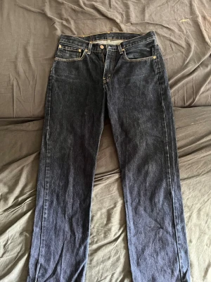 Mörkblå Levi's 501 jeans straight fit - Klassiska mörkblå Levi's 501 jeans med raka ben och fem fickor. Jeansen har gul kontrastsöm, knappgylf och den ikoniska läderpatchen bak i midjan. Tillverkade i slitstark denim, perfekt för en tidlös och avslappnad stil. Midja ish 36 innerben ish 84